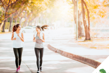 Zwei sportlich gekleidete Frauen joggen lachend nebeneinander auf einem von Bäumen gesäumten Weg in herbstlicher Atmosphäre. Die Sonne scheint durch das Laub, wodurch eine warme, einladende Stimmung entsteht.