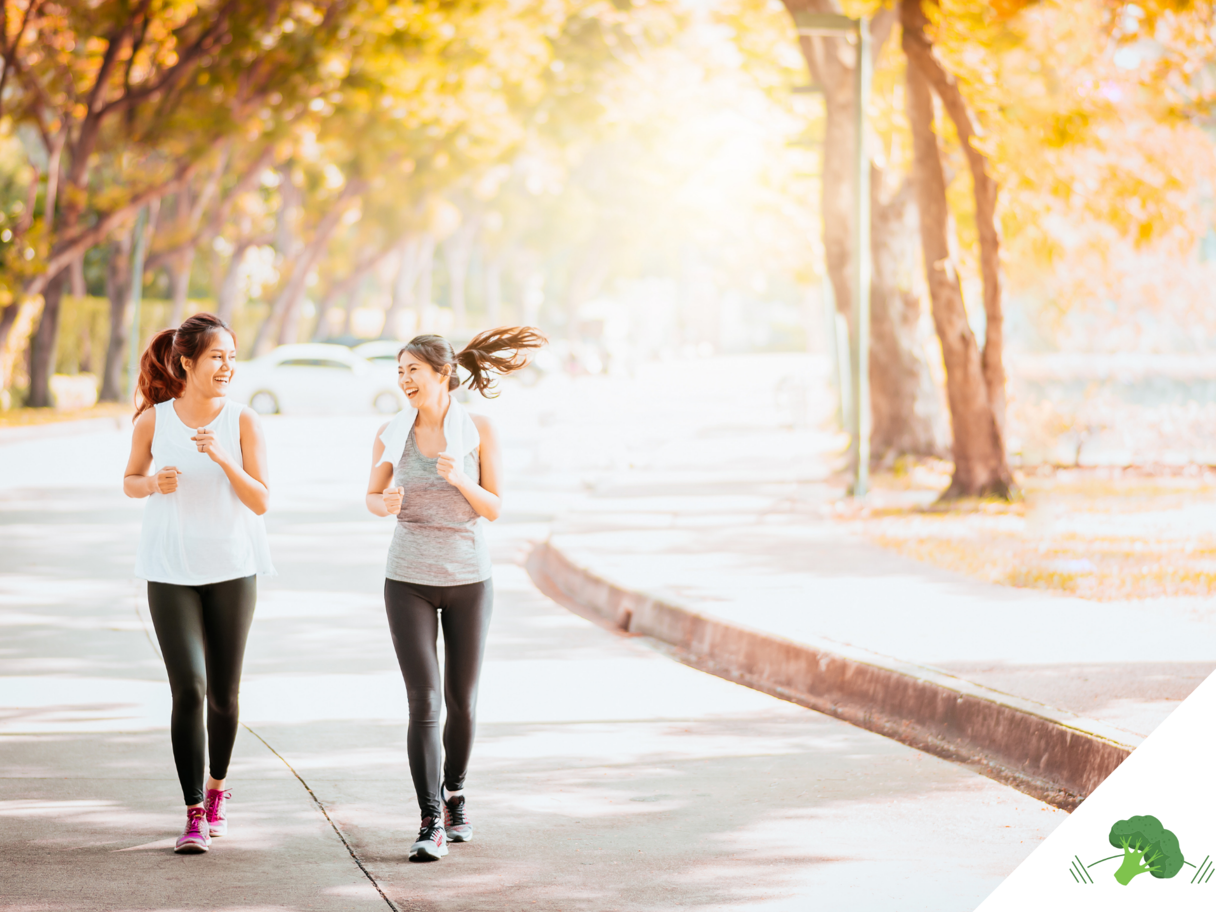 Zwei sportlich gekleidete Frauen joggen lachend nebeneinander auf einem von Bäumen gesäumten Weg in herbstlicher Atmosphäre. Die Sonne scheint durch das Laub, wodurch eine warme, einladende Stimmung entsteht.