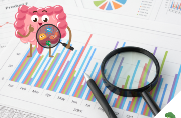 Fröhlich gezeichneter Darm mit Lupe betrachtet bunte Bakterien vor einem Hintergrund aus bunten Balken- und Kreisdiagrammen sowie Zahlen, daneben liegen eine echte Lupe und ein Stift auf einem Datenblatt.
