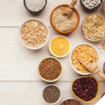 Schalen und Schüsseln mit verschiedenen pflanzlichen Lebensmitteln wie Linsen, Bohnen, Kichererbsen, Haferflocken, Chiasamen, Buchweizen und Getreide, stilvoll auf einem hellen Holztisch arrangiert, fotografiert aus der Vogelperspektive.