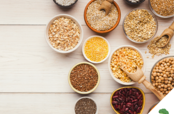 Schalen und Schüsseln mit verschiedenen pflanzlichen Lebensmitteln wie Linsen, Bohnen, Kichererbsen, Haferflocken, Chiasamen, Buchweizen und Getreide, stilvoll auf einem hellen Holztisch arrangiert, fotografiert aus der Vogelperspektive.