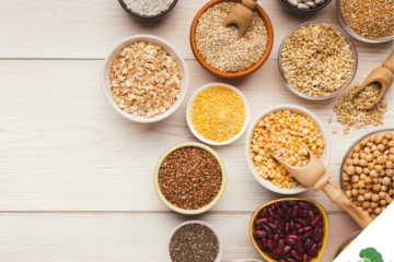 Schalen und Schüsseln mit verschiedenen pflanzlichen Lebensmitteln wie Linsen, Bohnen, Kichererbsen, Haferflocken, Chiasamen, Buchweizen und Getreide, stilvoll auf einem hellen Holztisch arrangiert, fotografiert aus der Vogelperspektive.