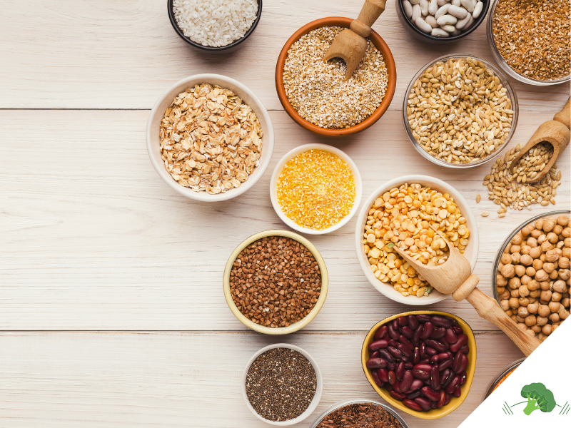 Schalen und Schüsseln mit verschiedenen pflanzlichen Lebensmitteln wie Linsen, Bohnen, Kichererbsen, Haferflocken, Chiasamen, Buchweizen und Getreide, stilvoll auf einem hellen Holztisch arrangiert, fotografiert aus der Vogelperspektive.