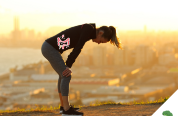 Sportlerin steht beim Laufen auf einem Hügel im Sonnenuntergang, nach vorne gebeugt mit den Händen auf den Knien abgestützt, während eine grafische Darstellung eines schmerzenden Darms im Bauchbereich eingeblendet ist; im Hintergrund ist eine Stadt unscharf zu sehen