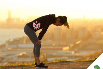 Sportlerin steht beim Laufen auf einem Hügel im Sonnenuntergang, nach vorne gebeugt mit den Händen auf den Knien abgestützt, während eine grafische Darstellung eines schmerzenden Darms im Bauchbereich eingeblendet ist; im Hintergrund ist eine Stadt unscharf zu sehen