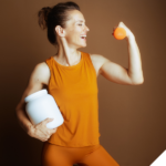 Sportliche Frau in orangefarbener Sportkleidung, die lächelnd ihren angespannten Bizeps zeigt, in der einen Hand einen Shaker hält und in der anderen eine Orange vor einem einfarbigen Hintergrund