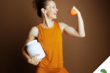 Sportliche Frau in orangefarbener Sportkleidung, die lächelnd ihren angespannten Bizeps zeigt, in der einen Hand einen Shaker hält und in der anderen eine Orange vor einem einfarbigen Hintergrund