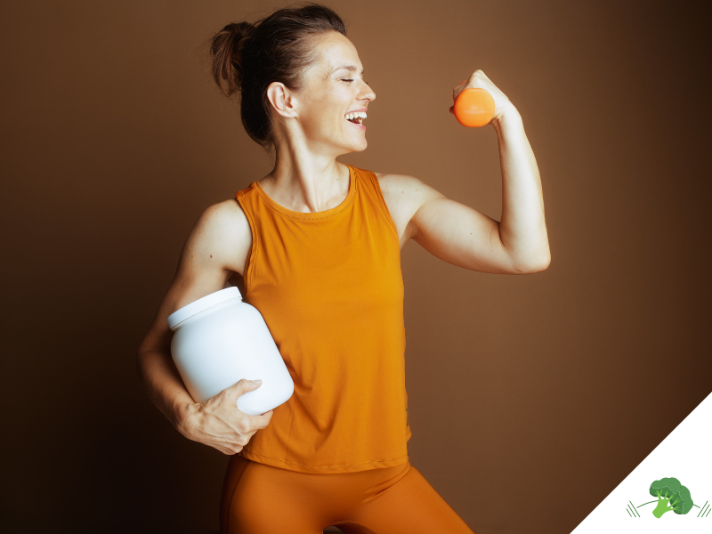 Sportliche Frau in orangefarbener Sportkleidung, die lächelnd ihren angespannten Bizeps zeigt, in der einen Hand einen Shaker hält und in der anderen eine Orange vor einem einfarbigen Hintergrund