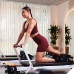 Frau in einem Pilates-Studio auf einem Reformer-Gerät in kniender Position, trägt ein bordeauxfarbenes Sportoutfit und führt eine Übung mit aufrechter Haltung aus, im Hintergrund helle Fenster und Pflanzen