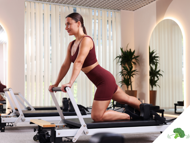 Frau in einem Pilates-Studio auf einem Reformer-Gerät in kniender Position, trägt ein bordeauxfarbenes Sportoutfit und führt eine Übung mit aufrechter Haltung aus, im Hintergrund helle Fenster und Pflanzen