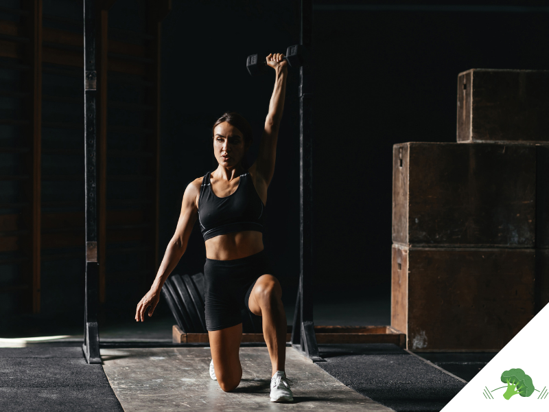 Frau in schwarzem Sport-Outfit macht in einer dunklen Trainingshalle einen Ausfallschritt und streckt eine Kurzhantel über den Kopf, im Hintergrund stehen Holzboxen und Hantelscheiben