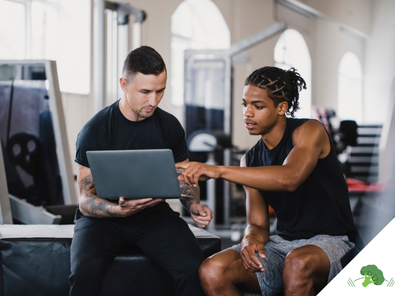 Trainer und junger Athlet sitzen im Fitnessstudio und schauen gemeinsam auf einen Laptop-Bildschirm, während der Athlet auf etwas zeigt