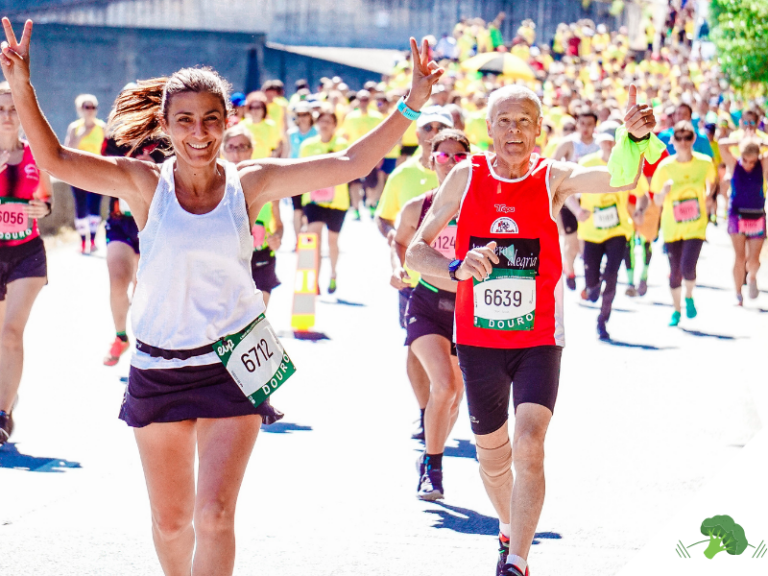 Lächelnde Läuferin mit erhobenen Armen und ein älterer Läufer mit Daumen hoch beim Marathon, umgeben von vielen weiteren Läufer:innen bei Sonnenschein