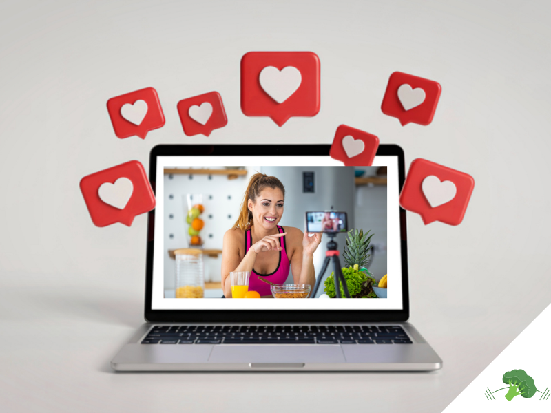Laptop mit einer lächelnden Frau im Sporttop, die in einer Küche vor einem Smartphone auf Stativ ein Ernährungsvideo aufnimmt, umgeben von roten Like-Symbolen mit Herzen